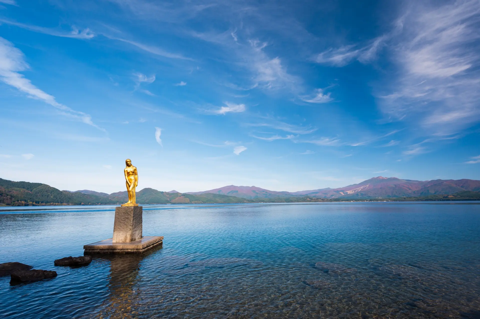Golden statue of a woman on a pedestal in a clear lake, with mountains under a blue sky.