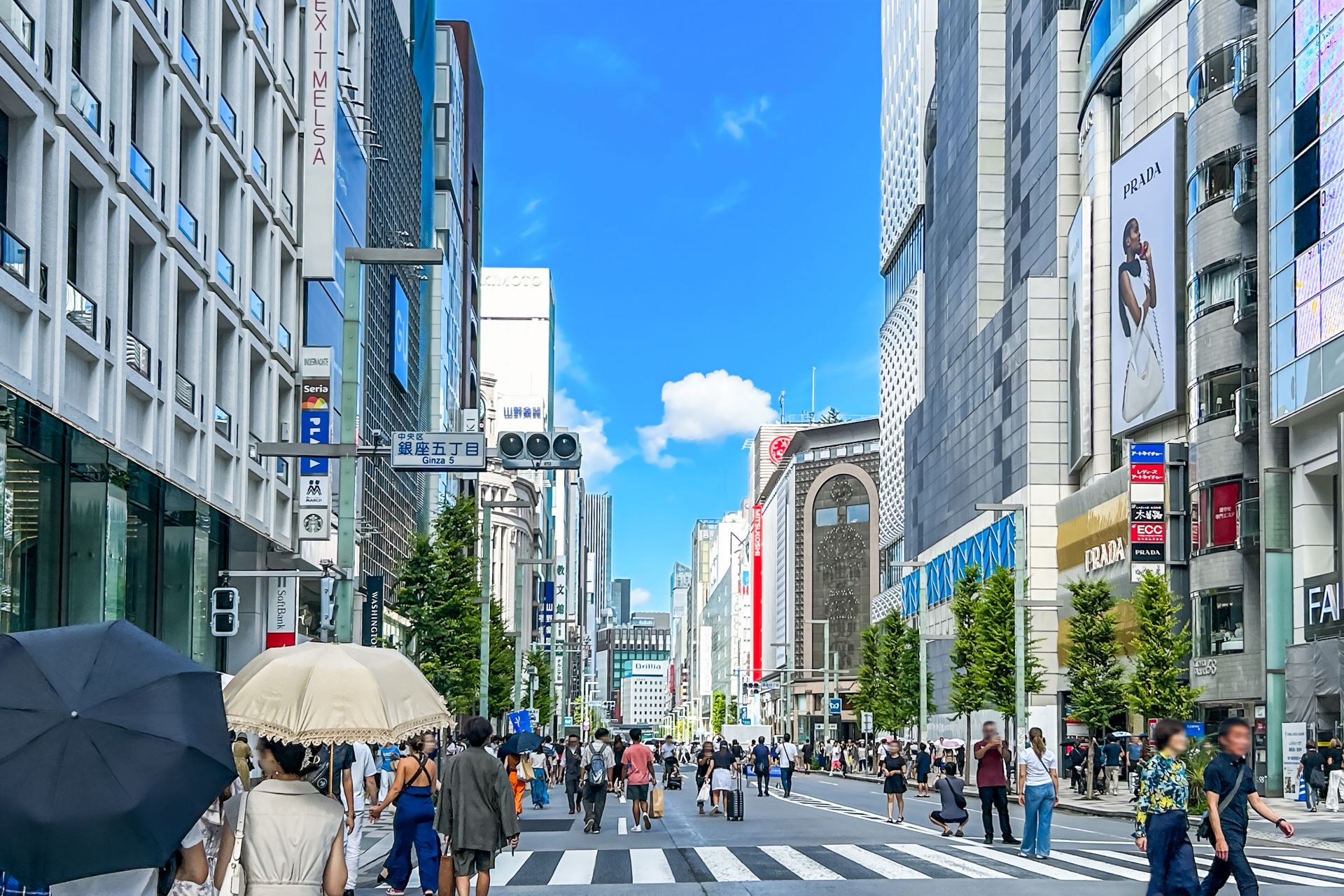 A bustling city street with pedestrians crossing a crosswalk, flanked by tall buildings with shops, under a clear blue sky.