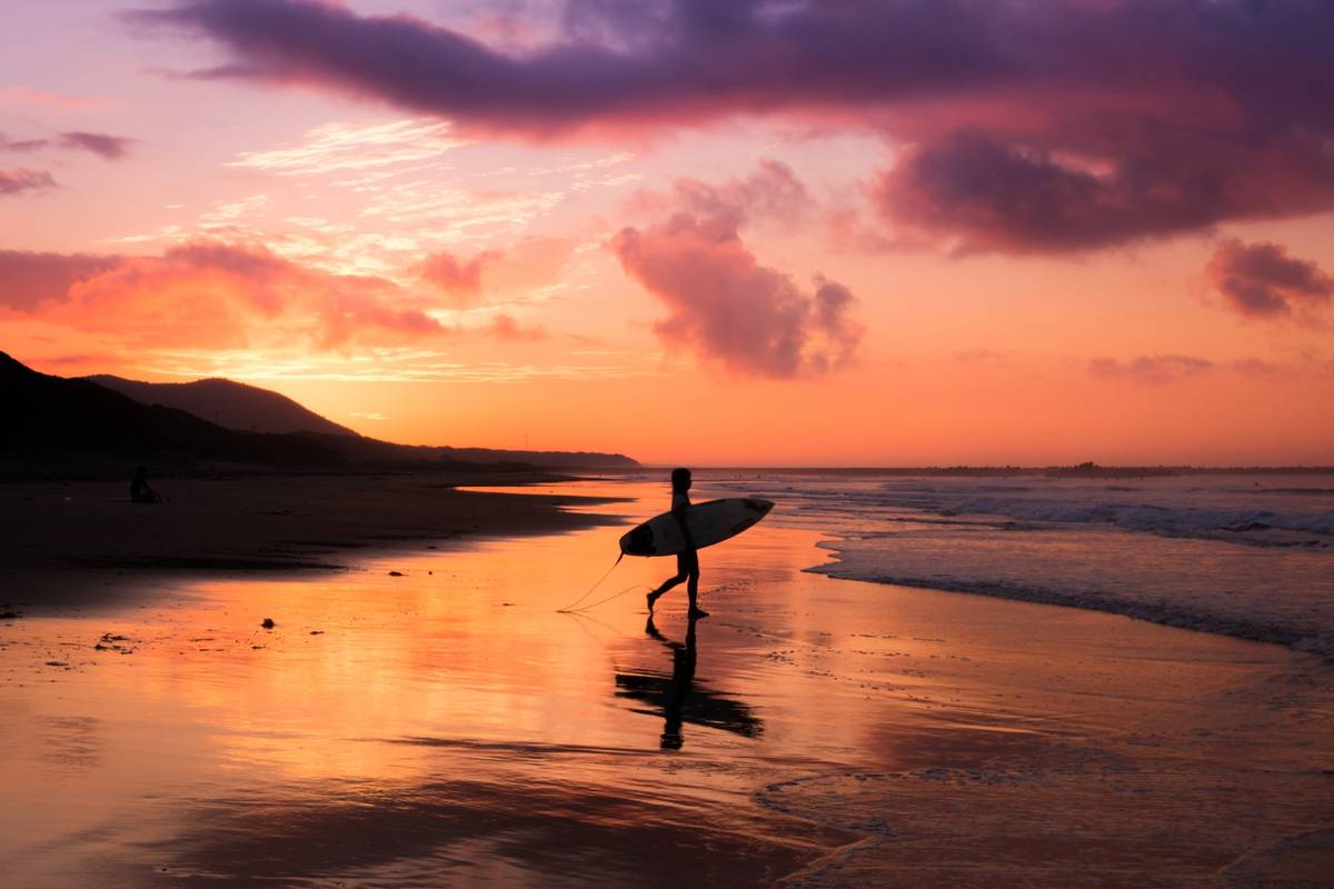 Silhouette of a surfer walking on a beach with a surfboard against a vibrant sunset.