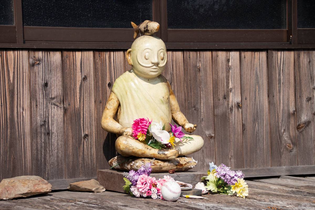 A meditating statue with a fish on its head, adorned with flowers and a baseball, against a wooden wall.