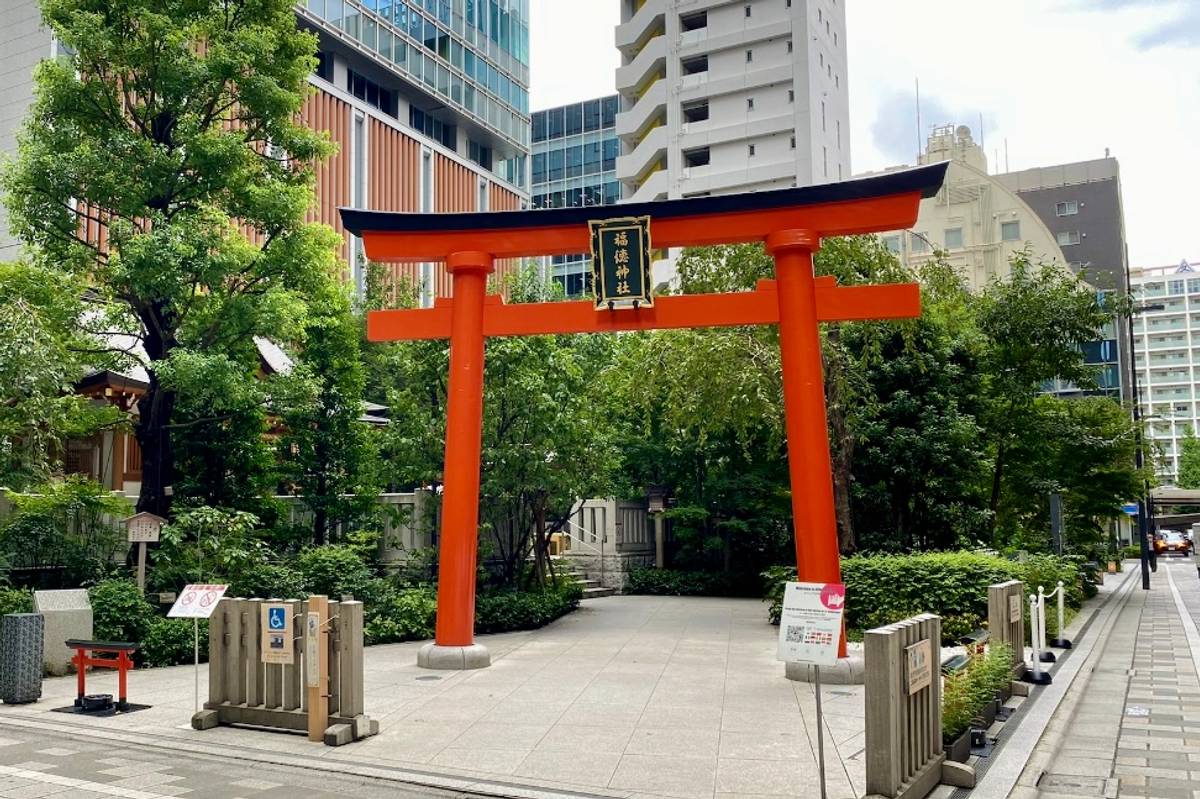 A vibrant orange torii gate stands in an urban park setting with lush green trees and modern buildings in the background.