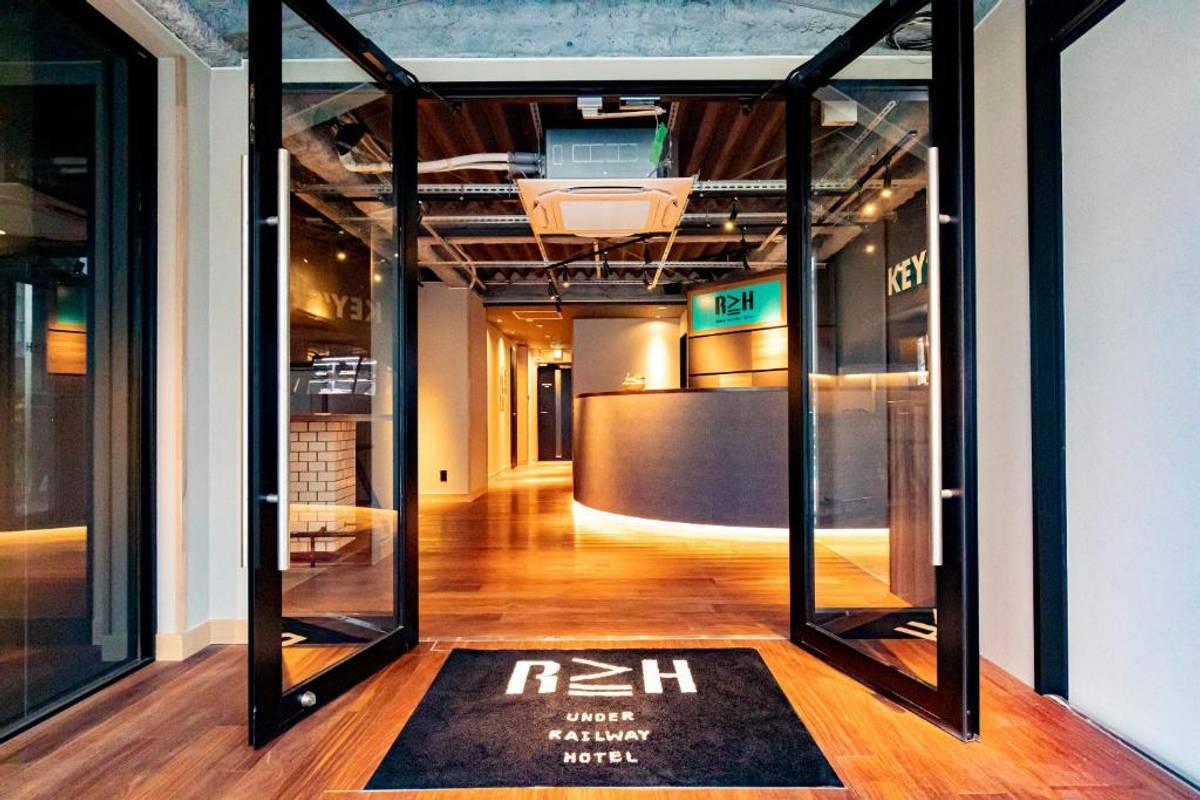 Modern hotel lobby seen through open glass doors with a curved reception desk, wooden floor, and exposed ceiling. A doormat reads 'R≥H UNDER RAILWAY HOTEL'.