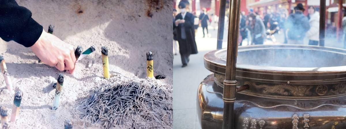 A hand places an incense stick into sand, next to a large, smoking communal incense burner at a temple.