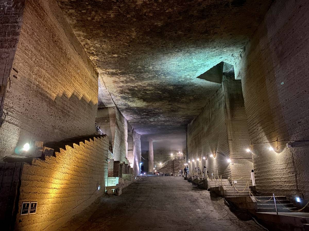 Vast underground quarry with rough-hewn walls and ceiling, illuminated by warm lights, featuring staircases and a wide path.