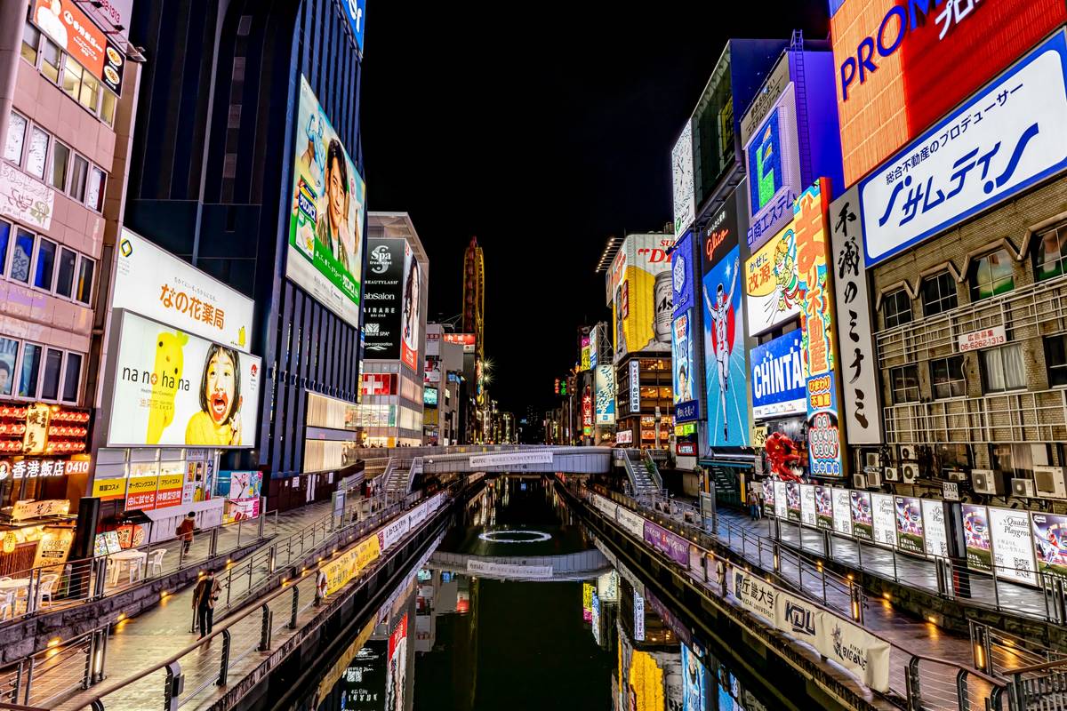 Vibrant night view of Osaka's Dotonbori canal, lined with brightly lit buildings and neon billboards, reflecting in the water.