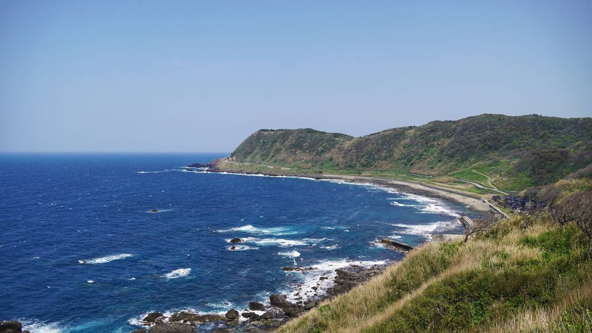 Coastal landscape with deep blue ocean, rocky shore, and green hills under a clear sky.