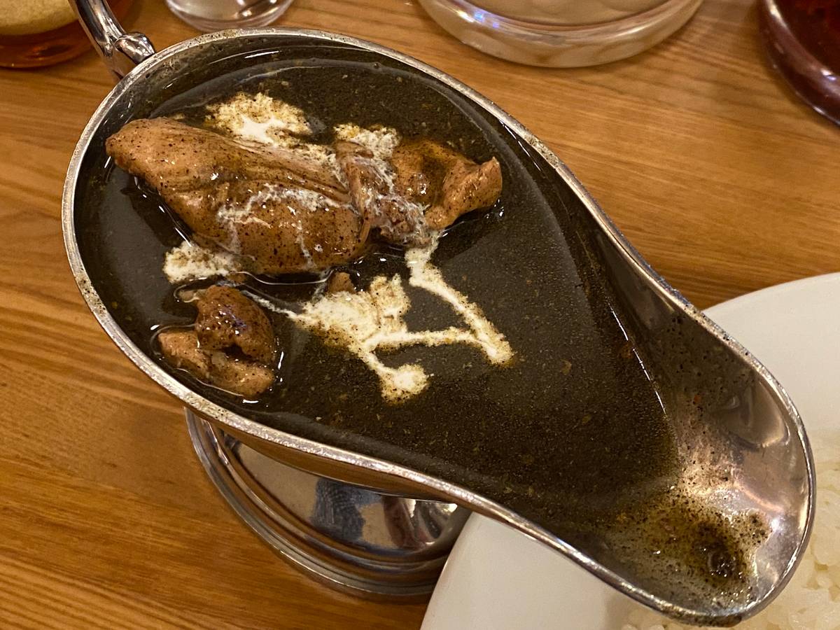 Dark chicken curry with white cream in a metal gravy boat on a wooden table.