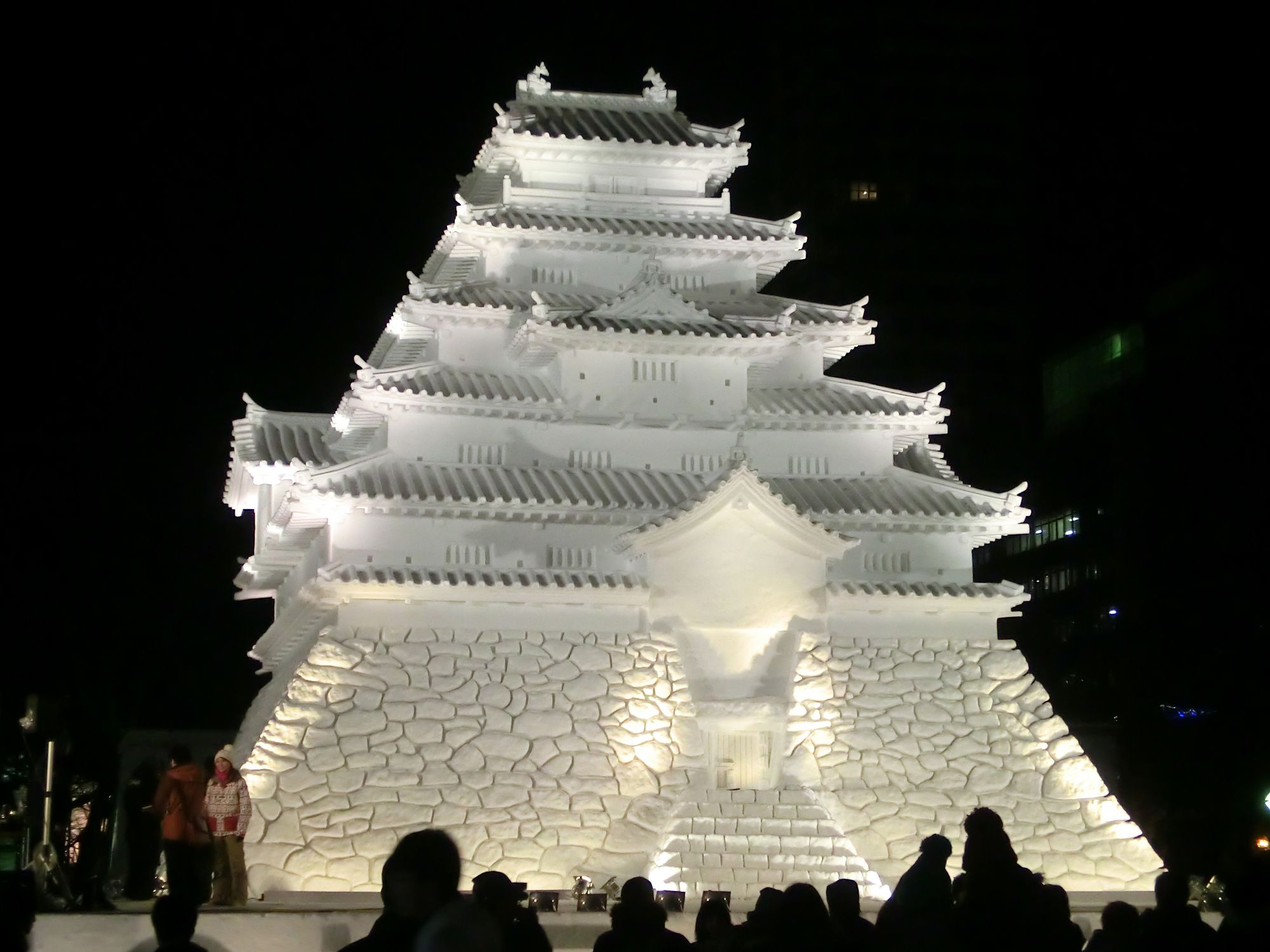 Illuminated snow sculpture of a multi-tiered Japanese castle at night, with a crowd gathered.