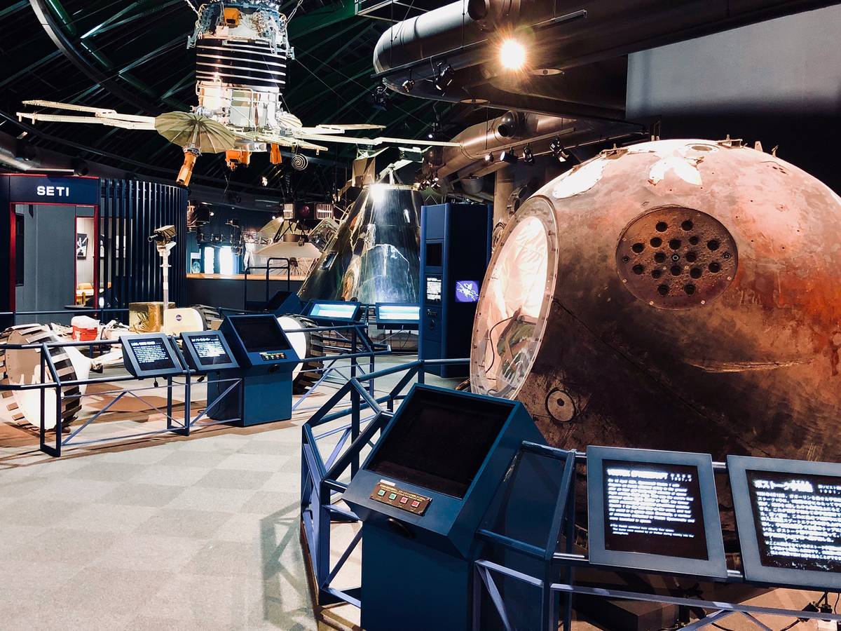 A space museum exhibition featuring a spherical spacecraft module, a hanging satellite, a rover, and interactive displays.