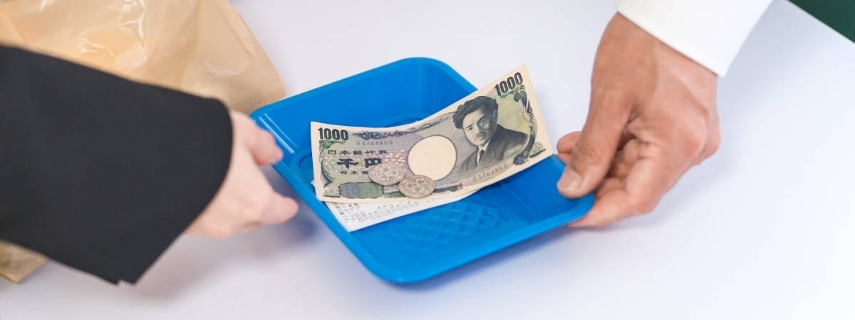Hands exchanging a blue tray holding a 1000 yen bill and coins.