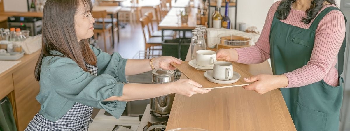Two women passing a tray with coffee cups across a cafe counter.