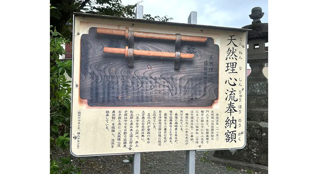 A wooden plaque dedicated to Ten'nen Rishin-ryū martial art, displaying two bokken and Japanese text, set outdoors.