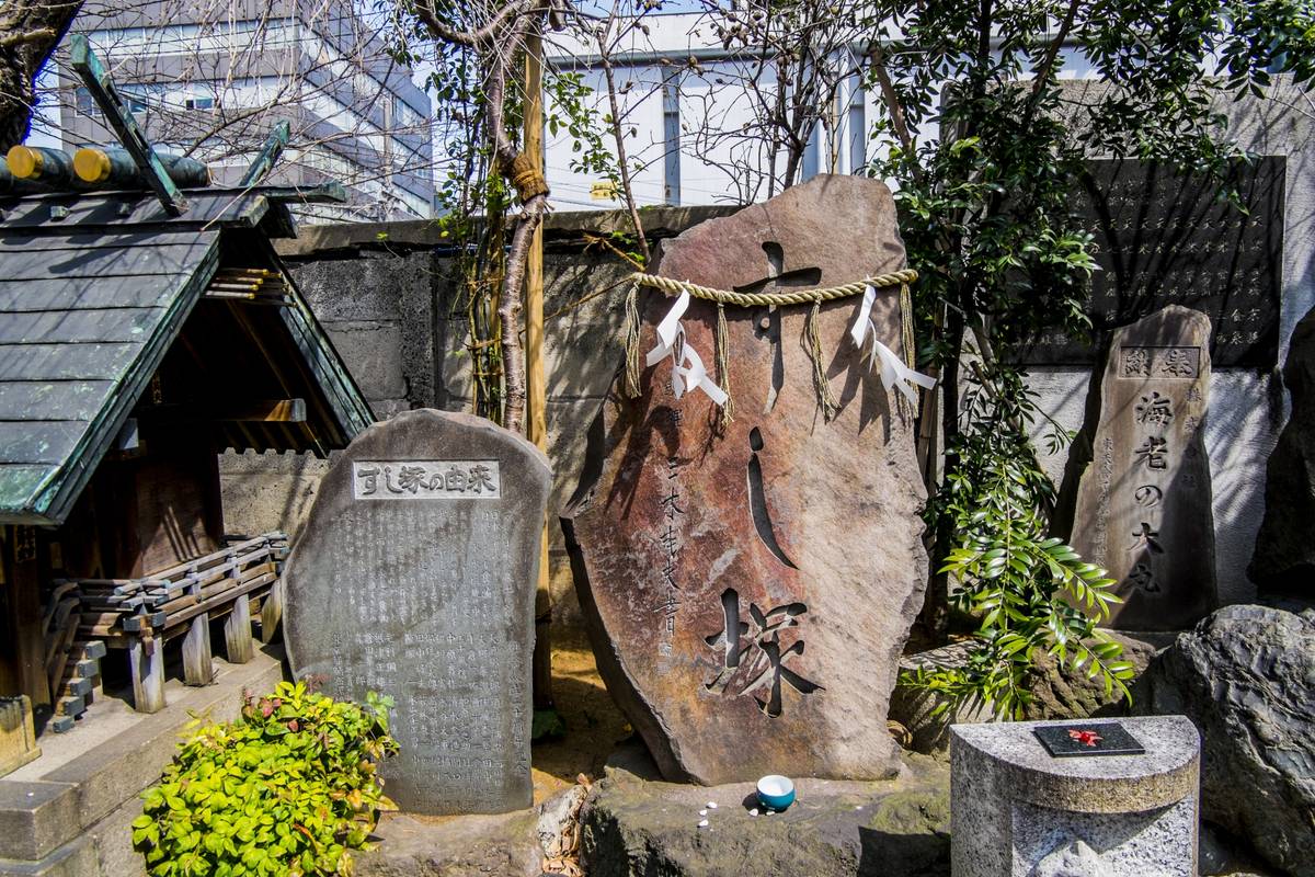 A small traditional Japanese shrine and several carved stone monuments, one decorated with a sacred rope and paper streamers, against a modern building in the background.