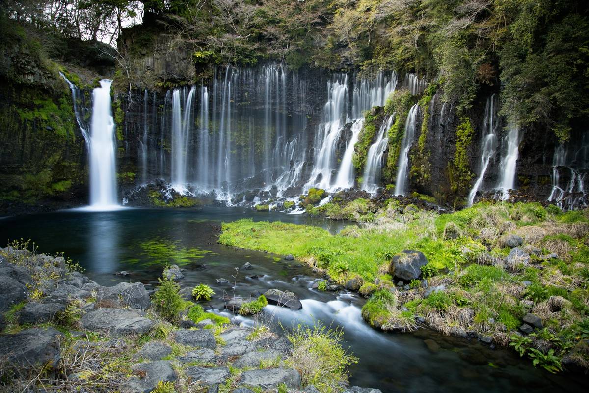 A wide curtain waterfall cascades down a mossy cliff into a dark pool, alongside a taller single waterfall on the left, surrounded by green vegetation and rocks.
