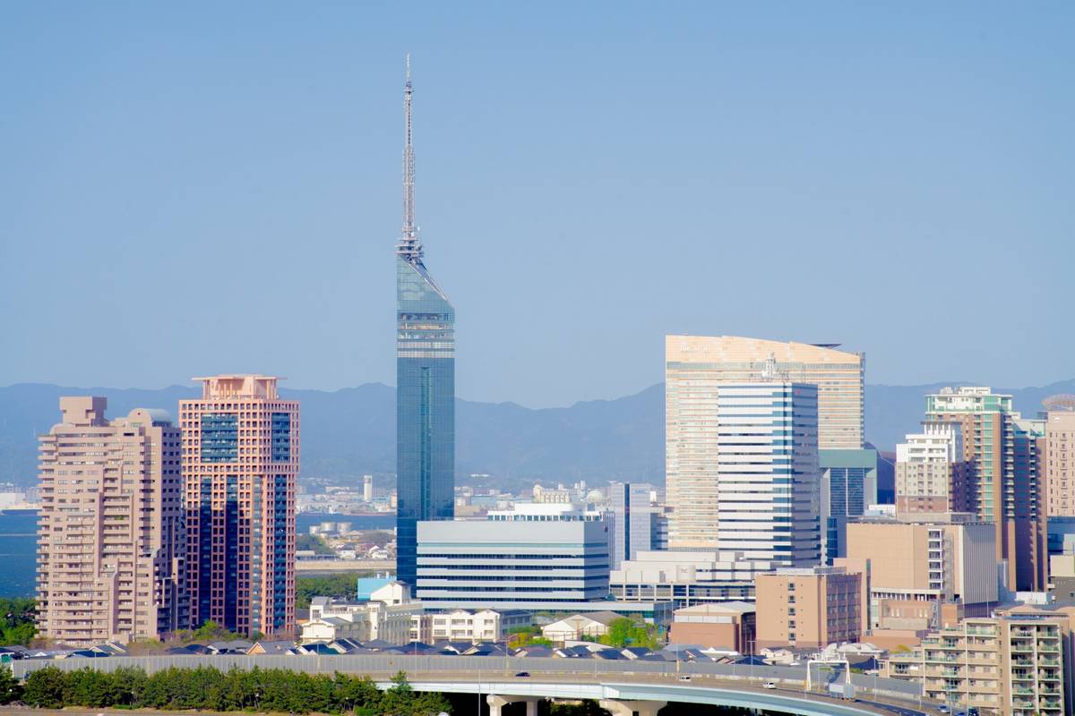 A tall blue glass skyscraper (Fukuoka Tower) stands prominently in a modern cityscape with other buildings, a clear blue sky, and distant mountains.