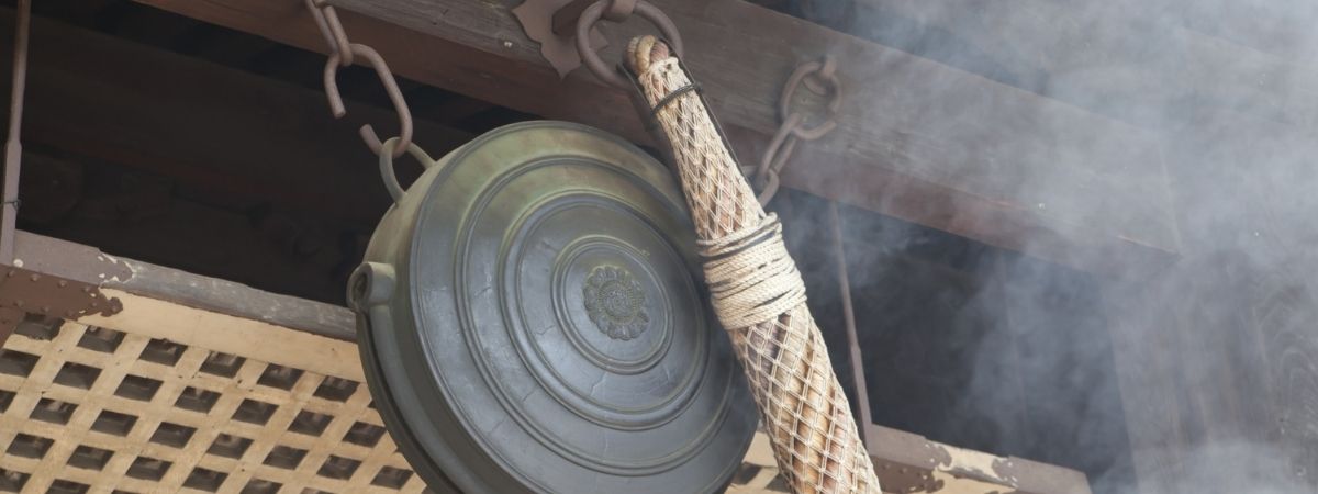A dark metal gong and a woven mallet hang from chains under a wooden structure, with smoke rising nearby.