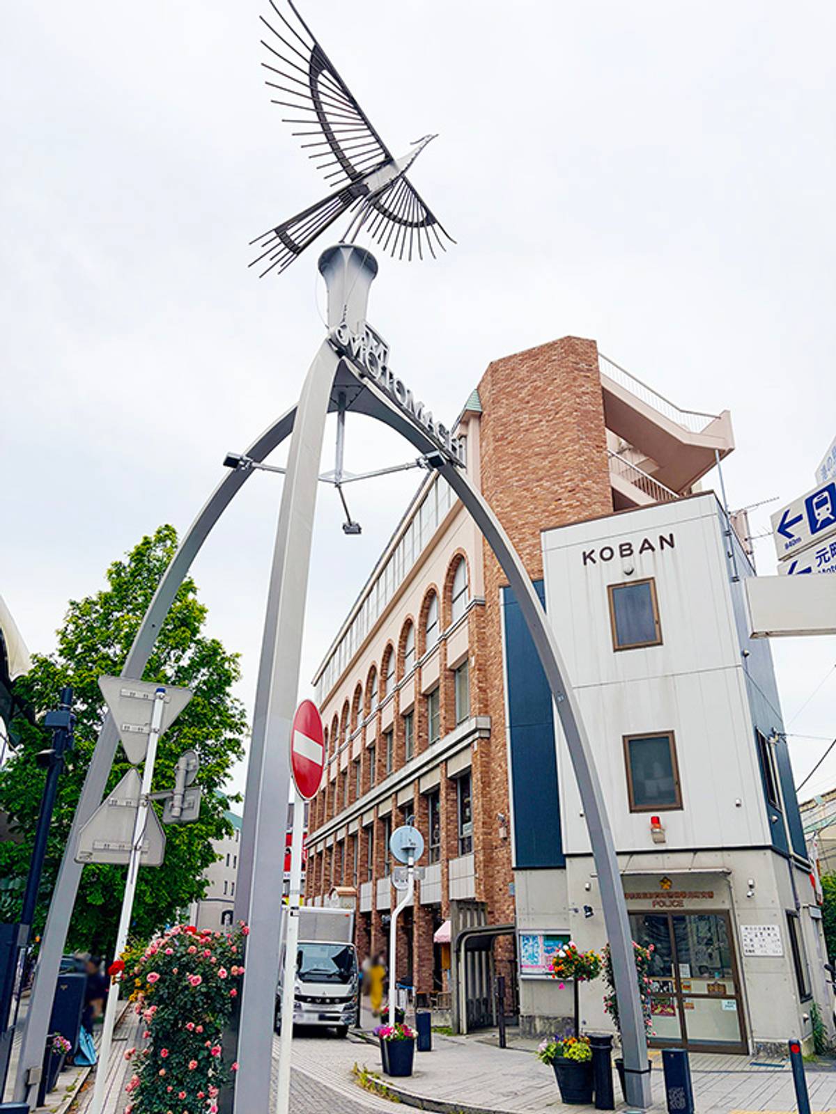 A street scene with a large gray archway topped by a metal bird sculpture, in front of a brick building and a white KOBAN police box.