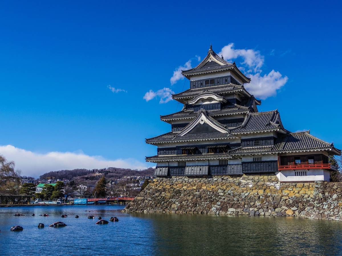 Dark tiered Japanese castle by a moat under a blue sky.