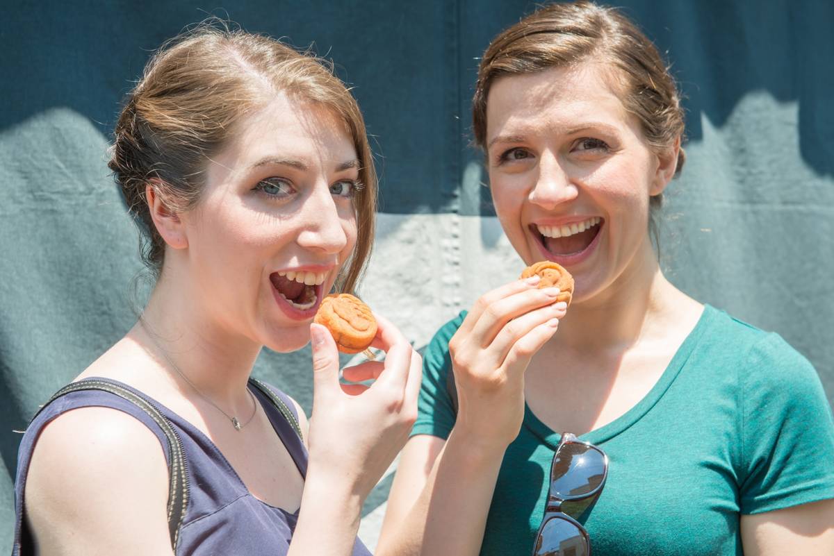 Two smiling women holding round cookies to their open mouths.