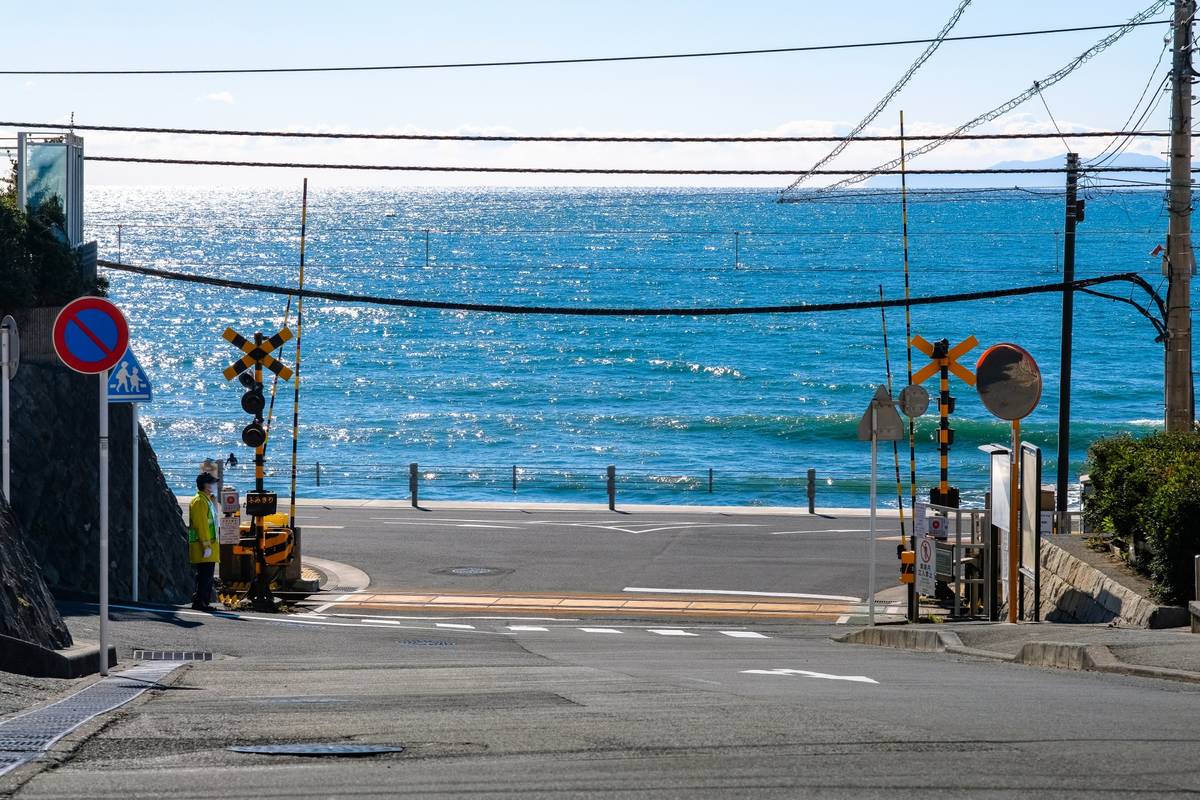 A road descends to a railroad crossing with two people, overlooking a shimmering blue ocean.
