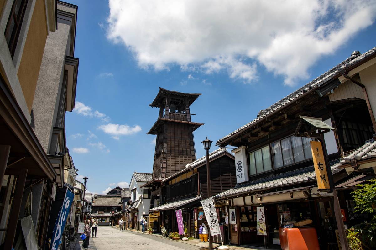 A historic Japanese street with traditional wooden buildings and a prominent bell tower under a blue sky.
