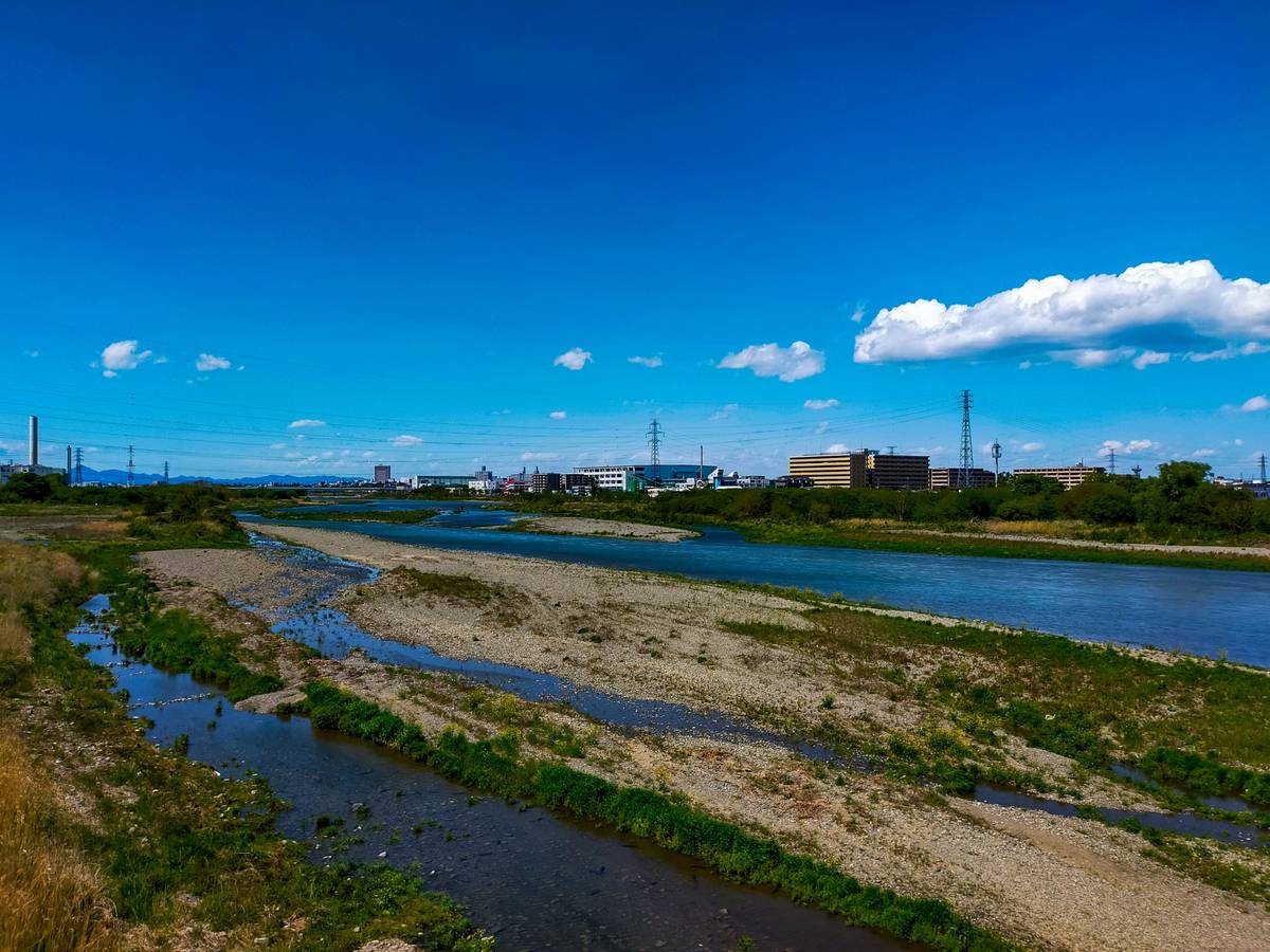 A wide riverbed with shallow water and gravel banks, industrial buildings, and power lines under a blue sky.
