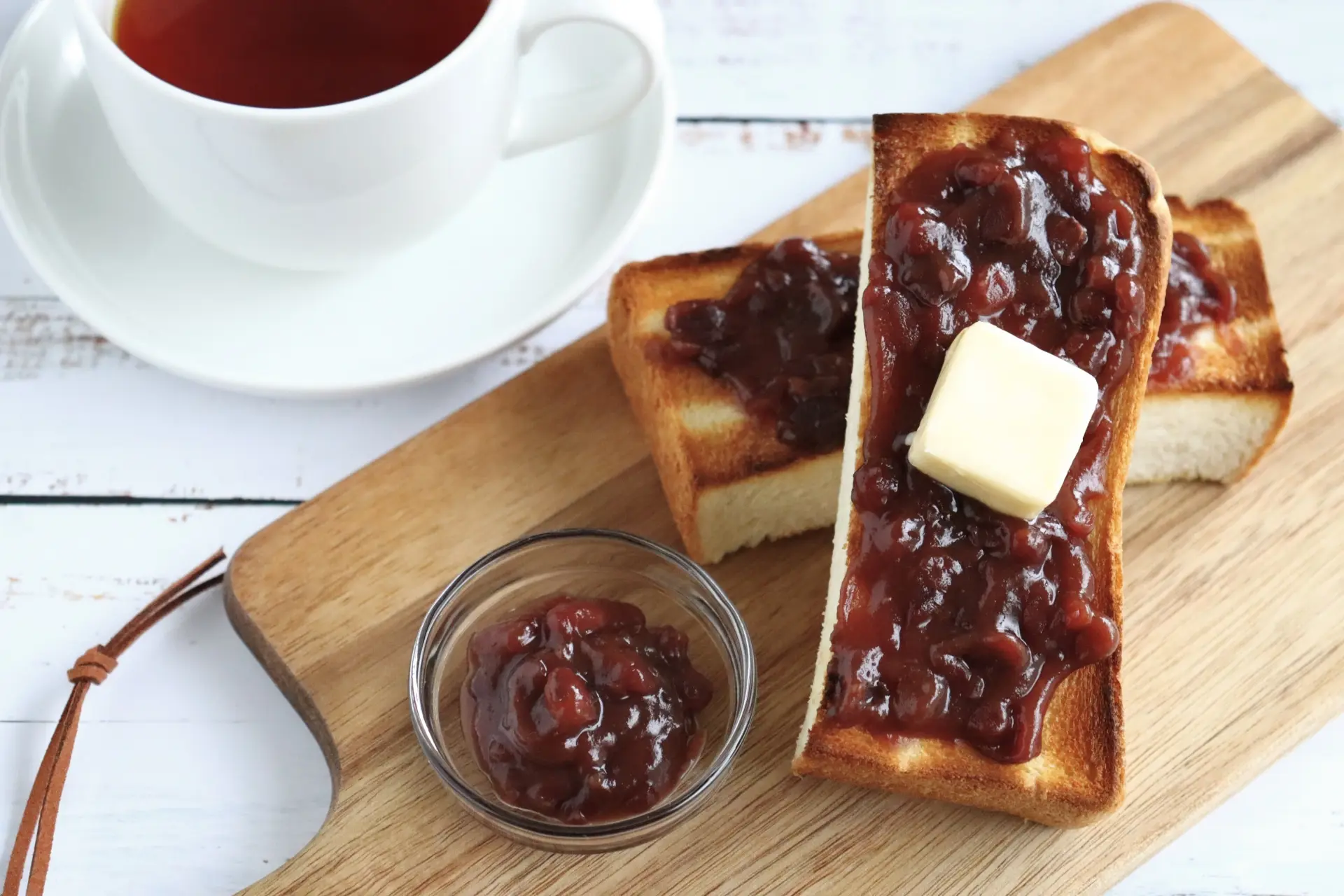 Two slices of toast topped with red bean paste, one with butter, alongside a cup of tea on a wooden board.