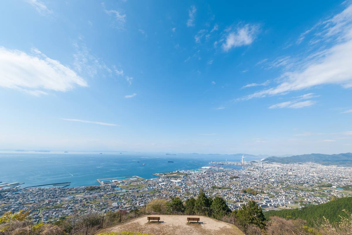 Elevated panoramic view of a coastal city, bay with ships, and distant mountains, with two benches in the foreground.