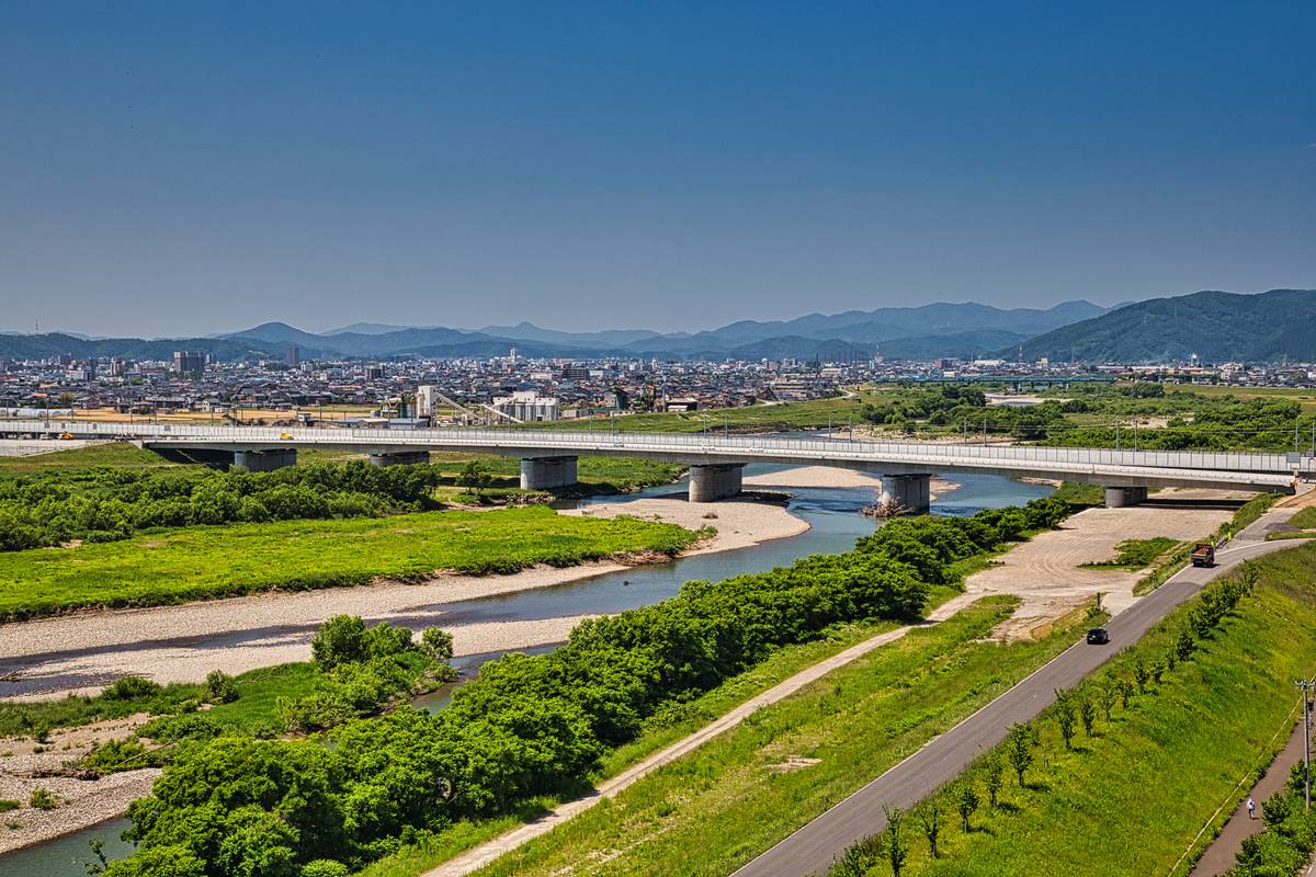 A long bridge spans a wide river with green banks, a road, a distant city, and mountains under a clear blue sky.