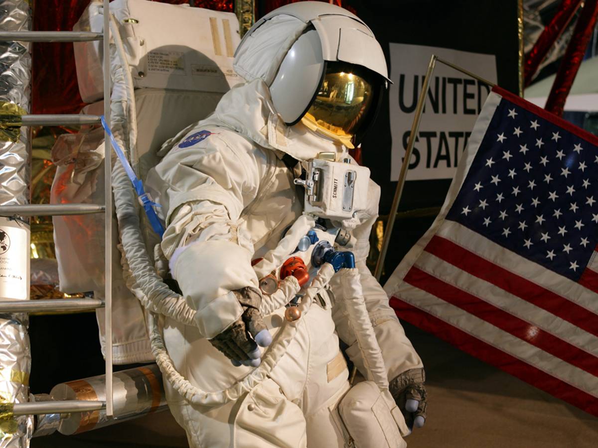 A white astronaut suit with a gold-visored helmet stands next to a United States flag.