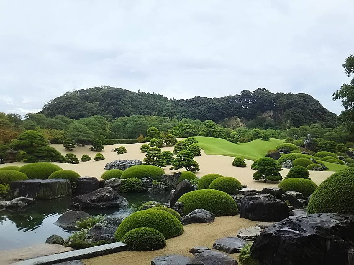 A Japanese garden with meticulously sculpted green mounds, raked sand, a pond, and pine trees, against forested hills.