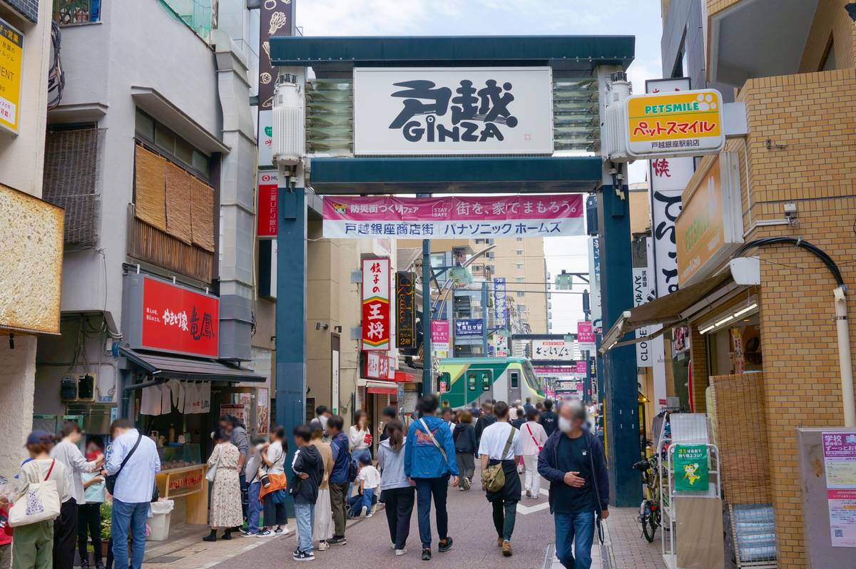 A busy street in Japan with an archway displaying "GINZA", numerous pedestrians, shops, and a train in the distance.