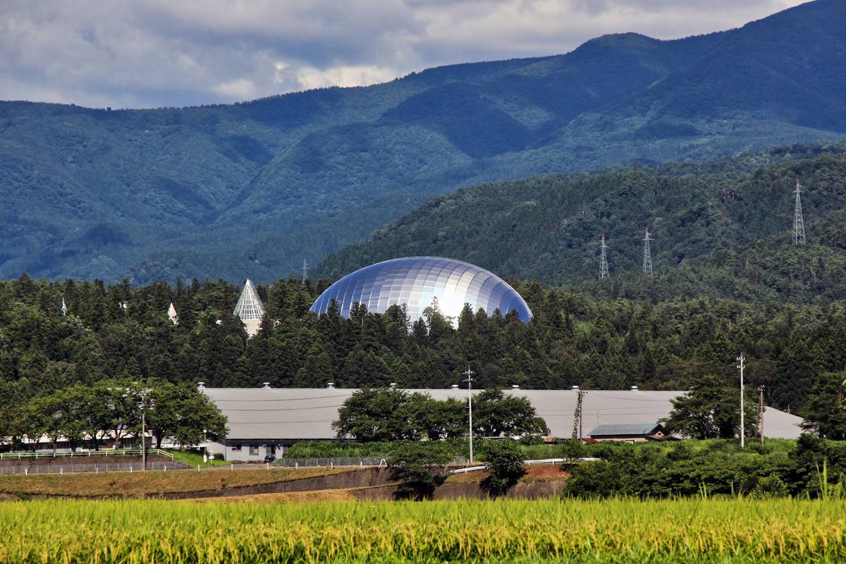 A large, reflective domed building surrounded by trees, with mountains in the background and a field of golden crops in the foreground.
