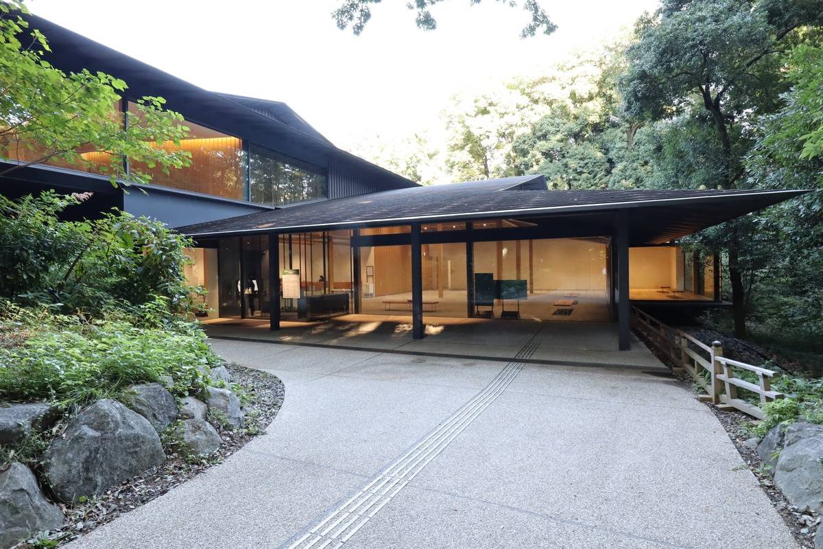 A modern building with a dark angular roof and glass walls, set amidst lush trees with a paved path.