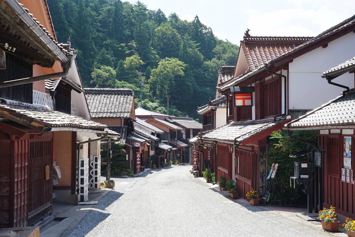 A traditional Japanese street lined with dark wood and white buildings with tiled roofs, backed by green mountains.