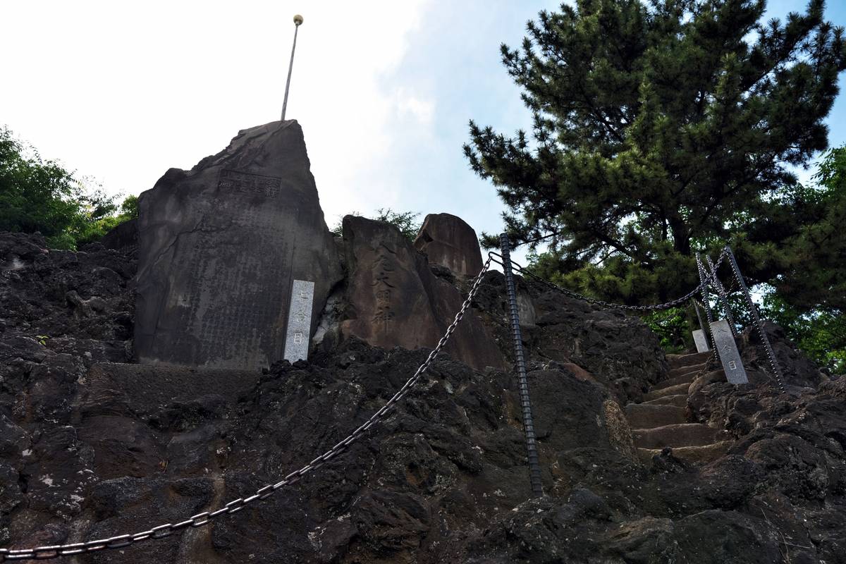 A rocky path with steps and chain railings leads up past a large inscribed rock monument topped with a golden orb, under a pine tree.
