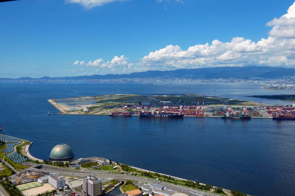 Aerial view of a container port in a bay, with a dome building on the near shore and a city with mountains in the distance.