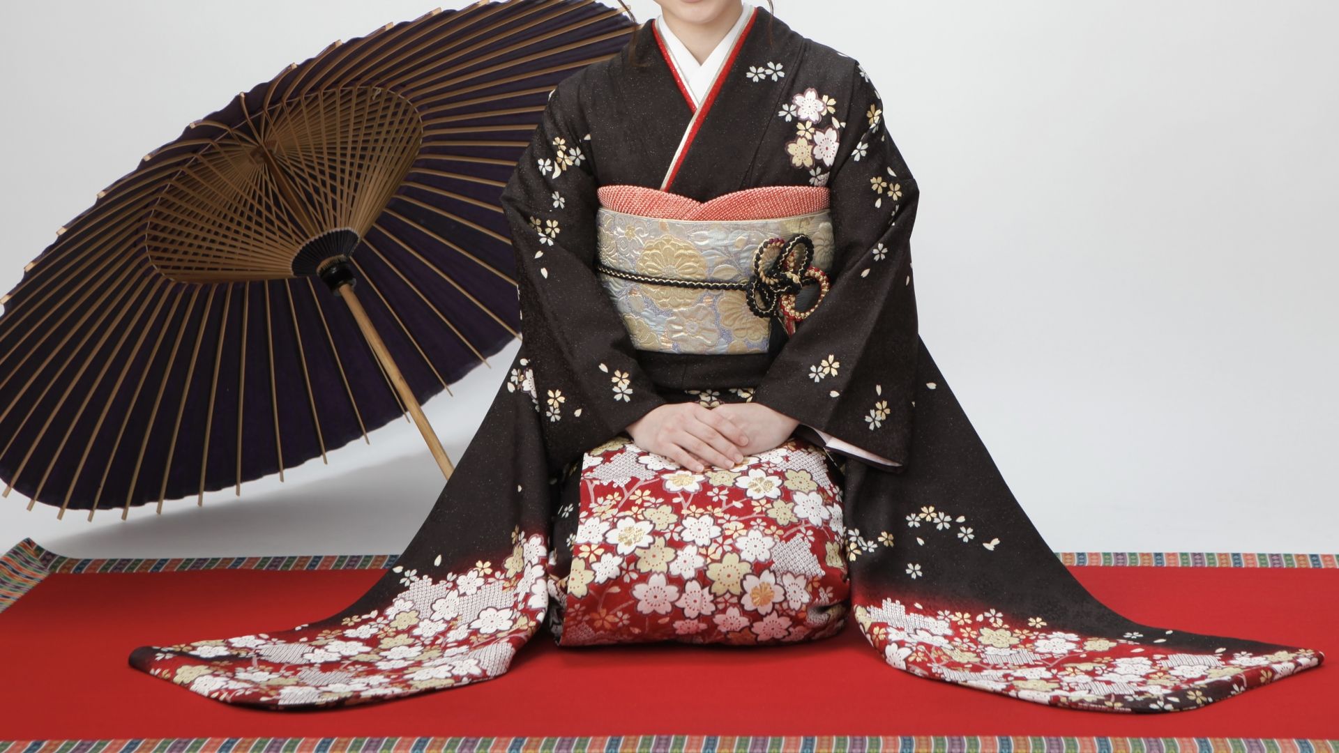 Kneeling person in a black floral kimono with an ornate obi, beside a traditional Japanese umbrella on a red mat.