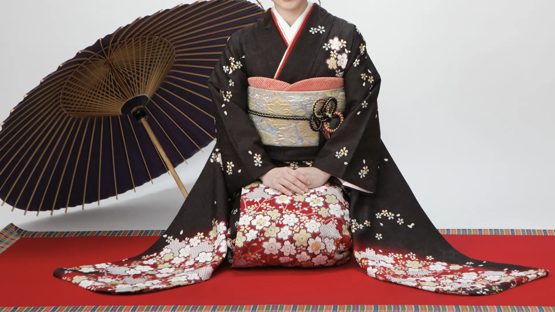 Kneeling person in a black floral kimono with an ornate obi, beside a traditional Japanese umbrella on a red mat.