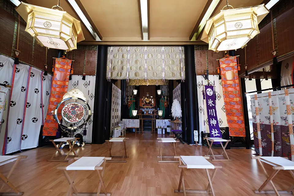 Interior of Hikawa Shinto Shrine with an altar, large lanterns, banners, a drum, and small stools.