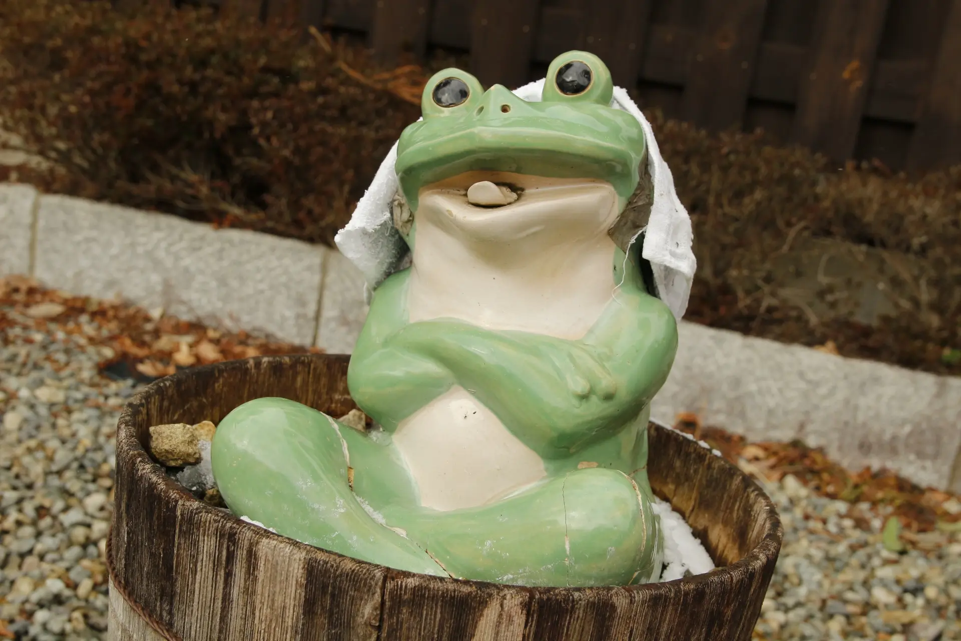 A green ceramic frog statue with a towel on its head, sitting in a wooden barrel.