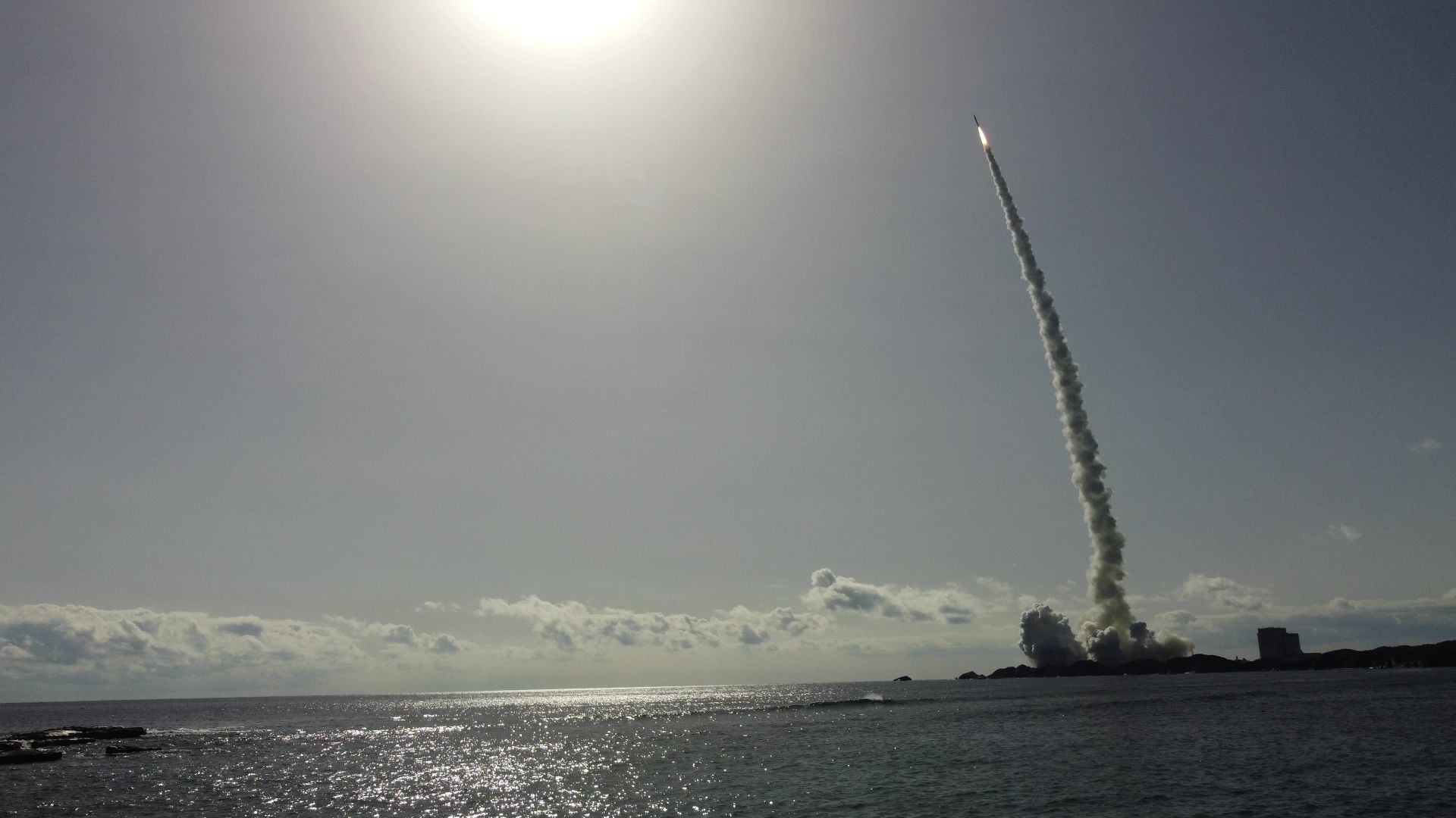 A rocket launching from a coastal area, leaving a trail of smoke, under a bright sun.