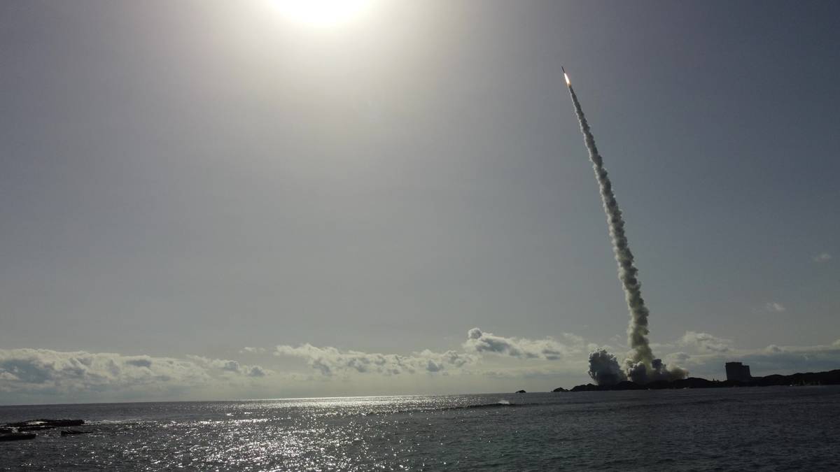 A rocket launching from a coastal area, leaving a trail of smoke, under a bright sun.