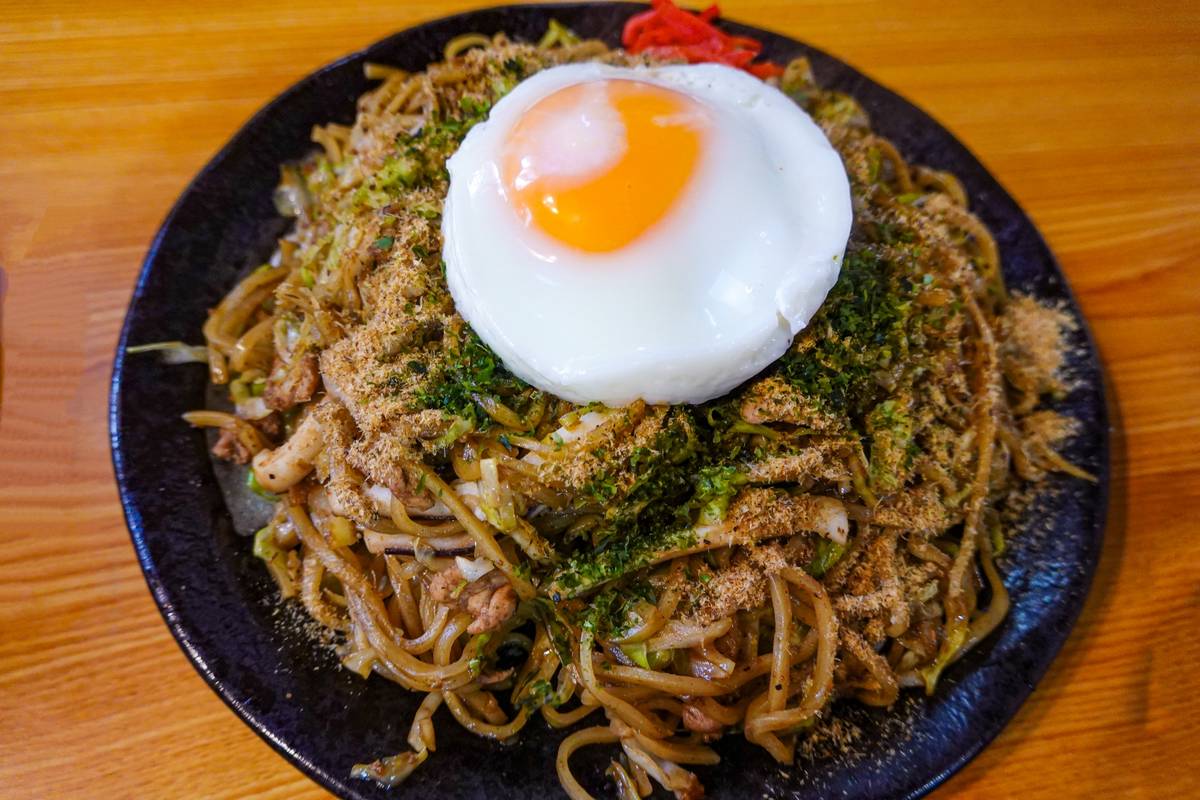 A plate of yakisoba noodles topped with a fried egg, nori, and bonito flakes.