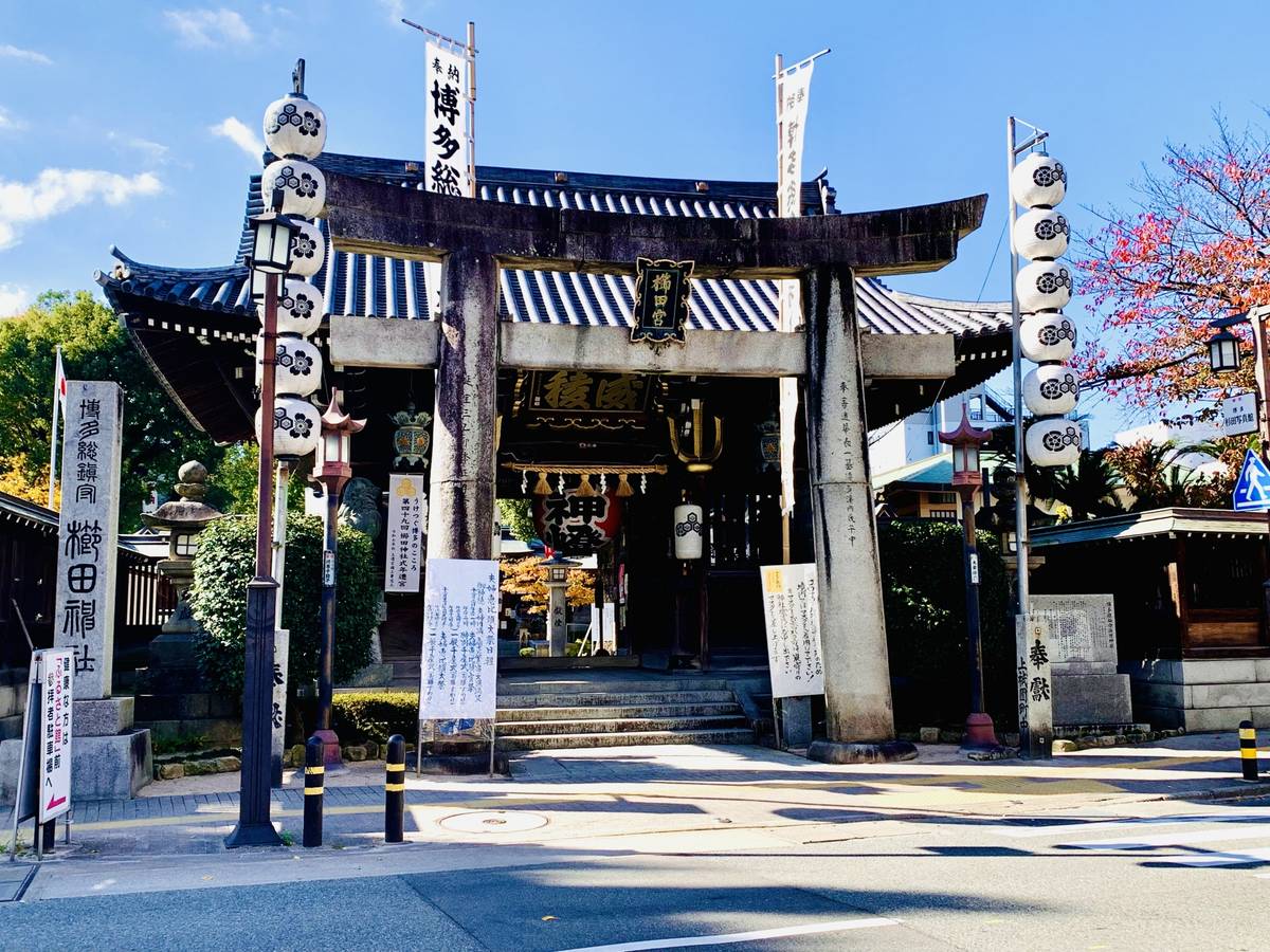 A stone Japanese temple gate with a tiled roof, flanked by tall poles holding stacked white lanterns and banners, under a blue sky.