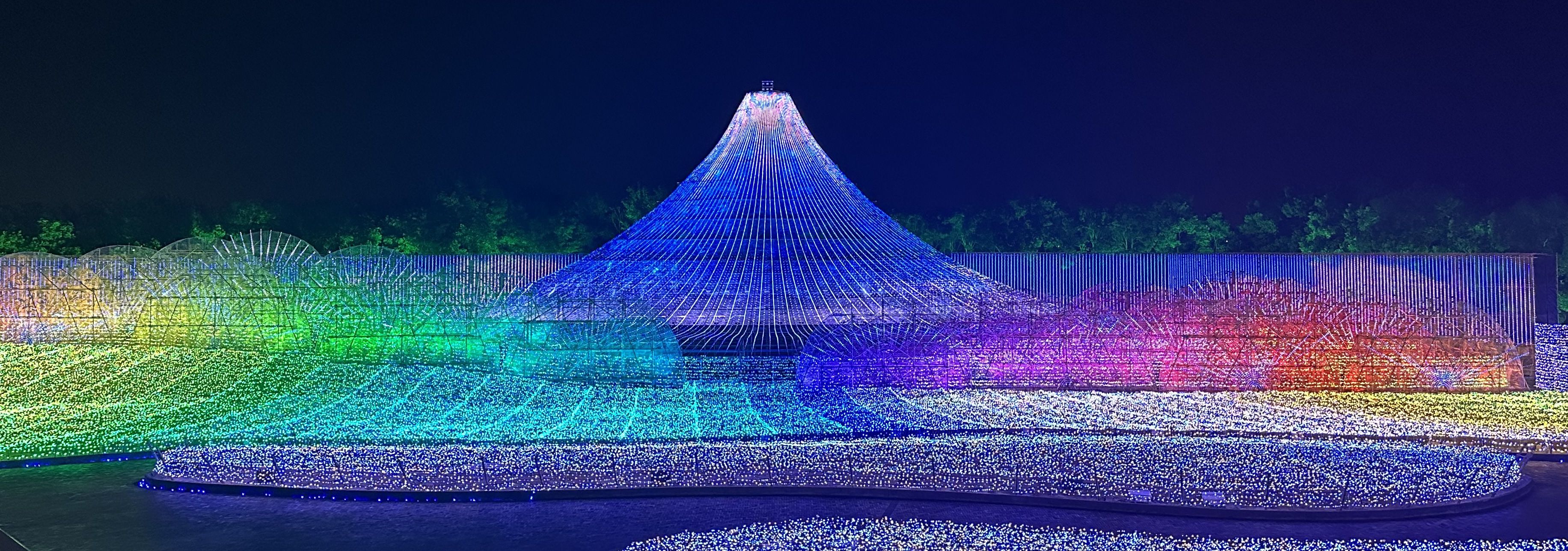 Nighttime light display featuring a blue conical Mount Fuji-like structure surrounded by waves of rainbow-colored lights.