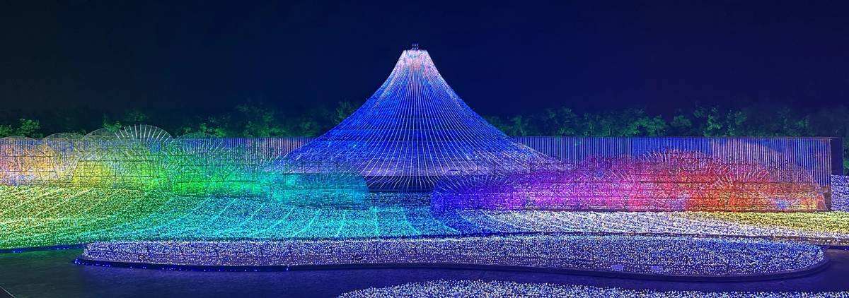 Nighttime light display featuring a blue conical Mount Fuji-like structure surrounded by waves of rainbow-colored lights.