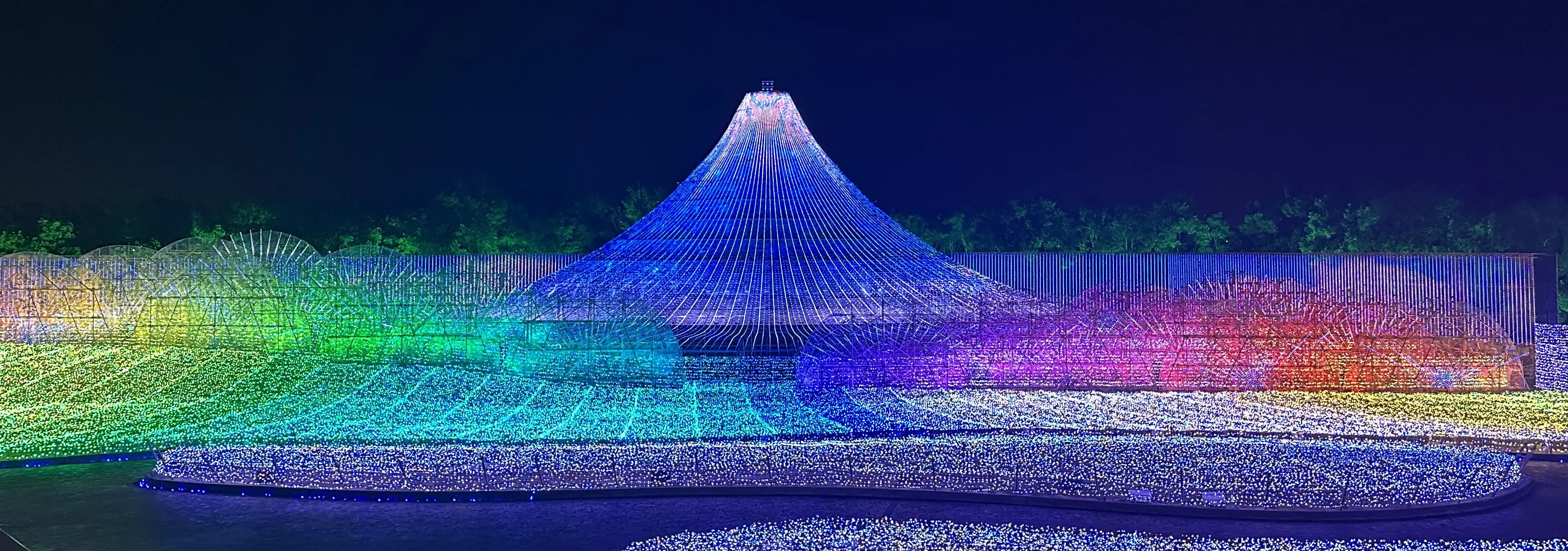 Nighttime light display featuring a blue conical Mount Fuji-like structure surrounded by waves of rainbow-colored lights.