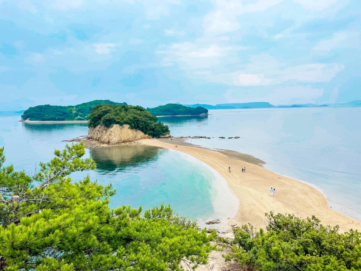 A long, narrow sandbar curves across calm turquoise water towards a small, tree-covered island. People walk on the sand.