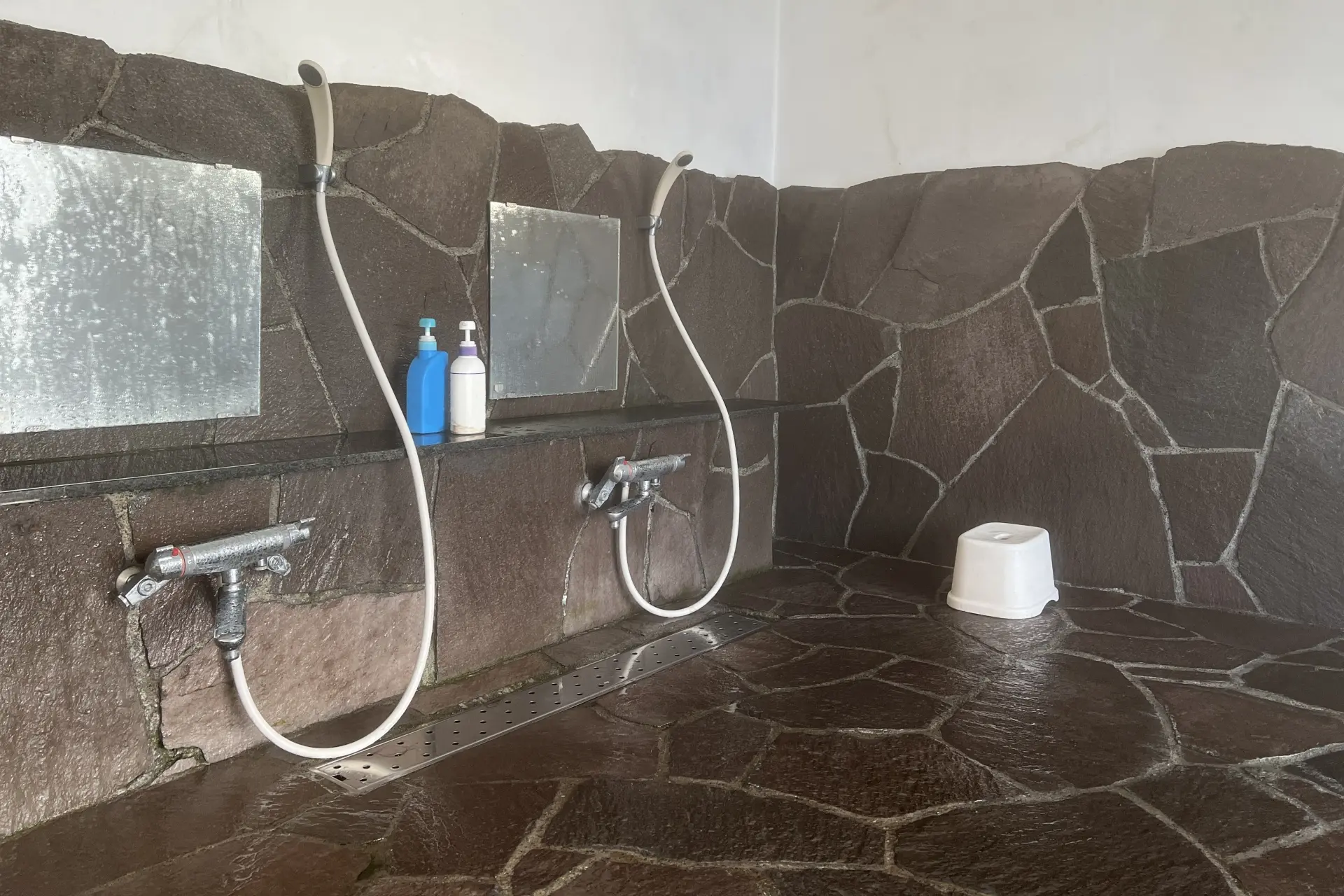 Communal shower area with dark irregular stone tiles, two showerheads, mirrors, toiletries, and a white stool.
